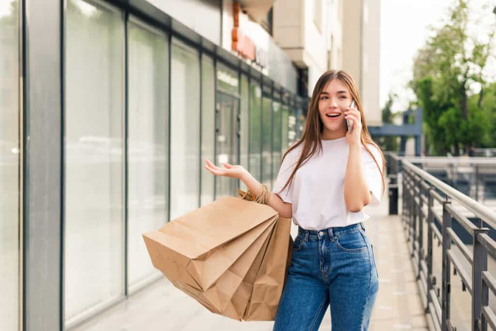 Einkaufen A young woman with long brown hair, wearing a white t-shirt and blue jeans, is walking outdoors past a modern storefront or apartment building. She is talking on a mobile phone, holding several large brown paper shopping bags in one arm, and gesturing with her free hand. She appears excited or engaged in the conversation.