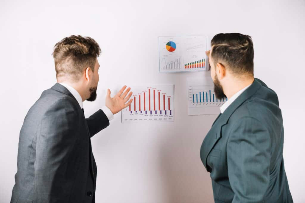 ETFs vs Stocks two business professionals standing in an office analyzing financial charts on a wall, pointing at bar and line graphs that compare market performance and investment trends.