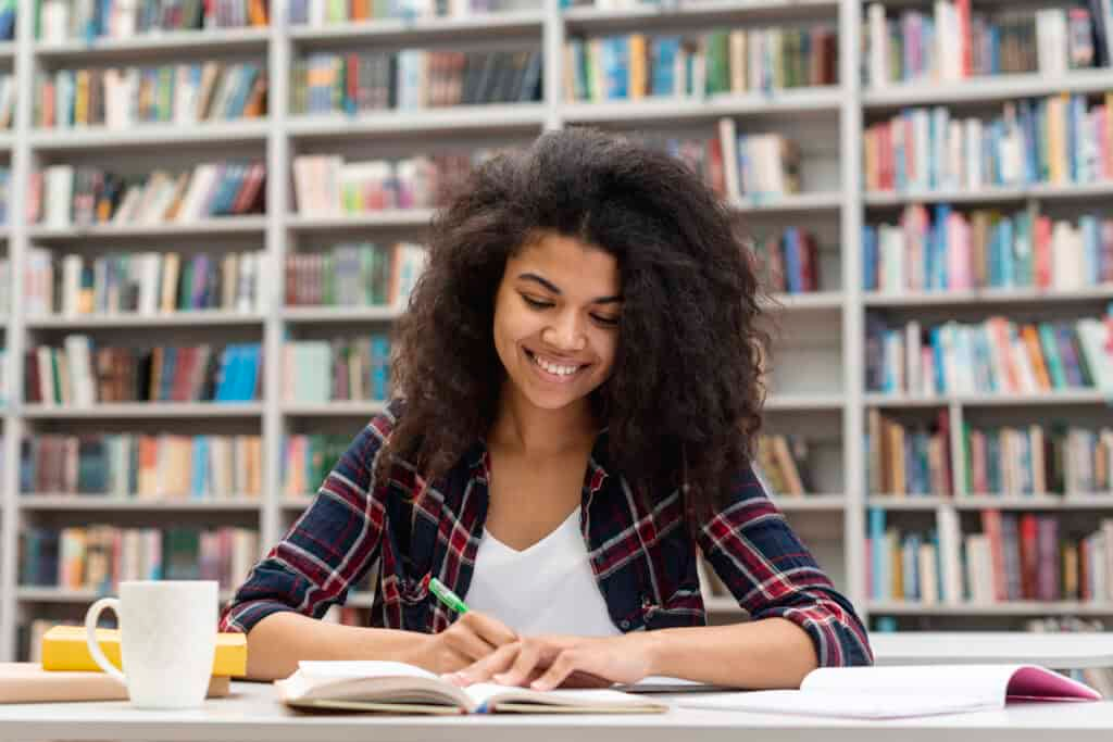 Reiseplanung Freelancing A young woman with curly dark hair and a bright smile sits at a desk, studying or reading a book. She is wearing a plaid shirt over a white top. The background is blurred but clearly shows tall bookshelves packed with colorful books, indicating she is in a library or bookstore.