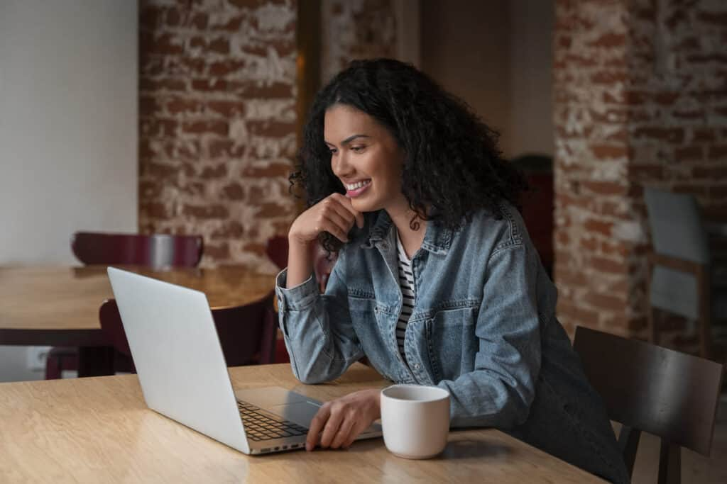 schufa The image features a young woman smiling warmly while engaging with a laptop in a cozy, brick-walled setting, likely a cafe or a co-working space.