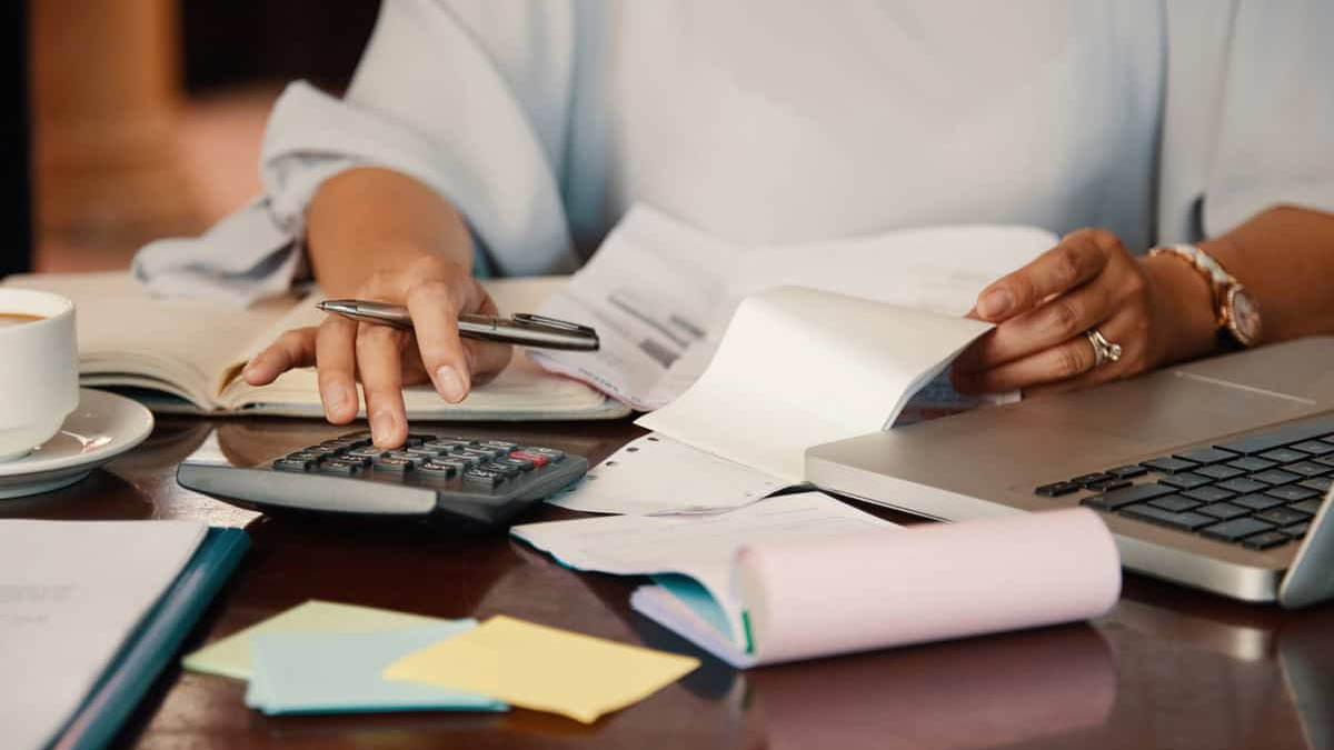 A close-up view of a person wearing a light-colored blouse, sitting at a desk and actively working on finances. One hand is using a calculator, while the other hand holds a document or receipt, suggesting they are managing a budget or calculating expenses. A notebook, a pen, and a white coffee cup are also visible on the desk.