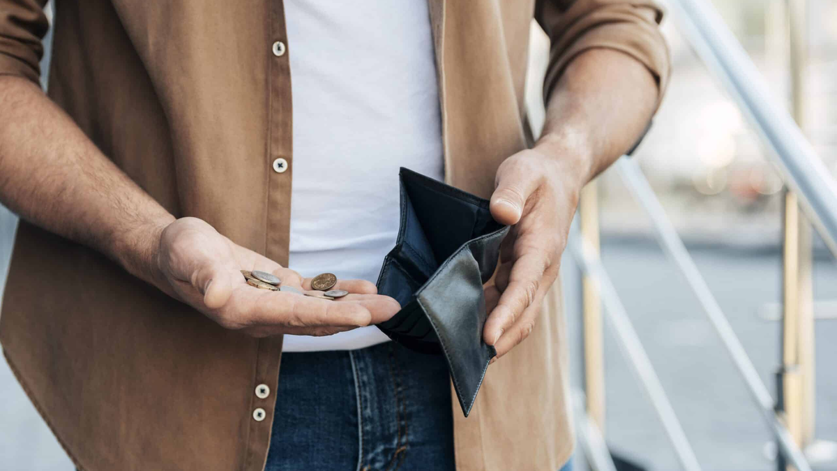 The image is a close-up of a person, likely a man, holding an empty black bifold wallet in one hand and a few small coins in the other.
