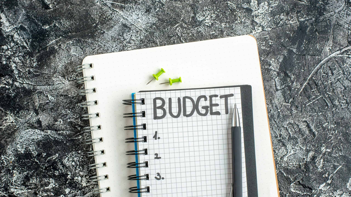 A small, grid-lined notepad with the word "BUDGET" written clearly on the top line, resting on a larger white notebook. A black pen and two small green thumbtacks are placed near the top of the notepad. The background is a dark, textured surface, providing a dramatic contrast to the white paper.
