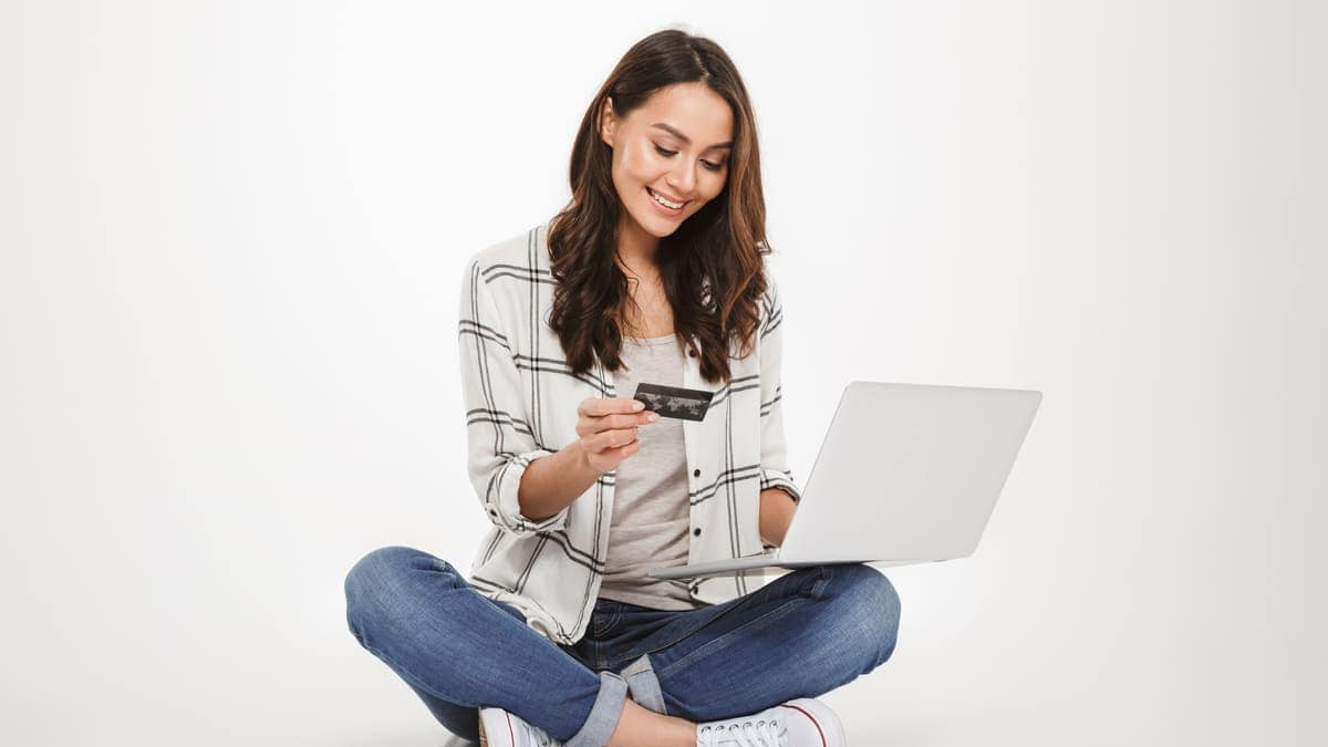 Budget The image is a studio shot of a young woman sitting cross-legged on the floor, using a laptop and a credit card, likely for an online transaction.
