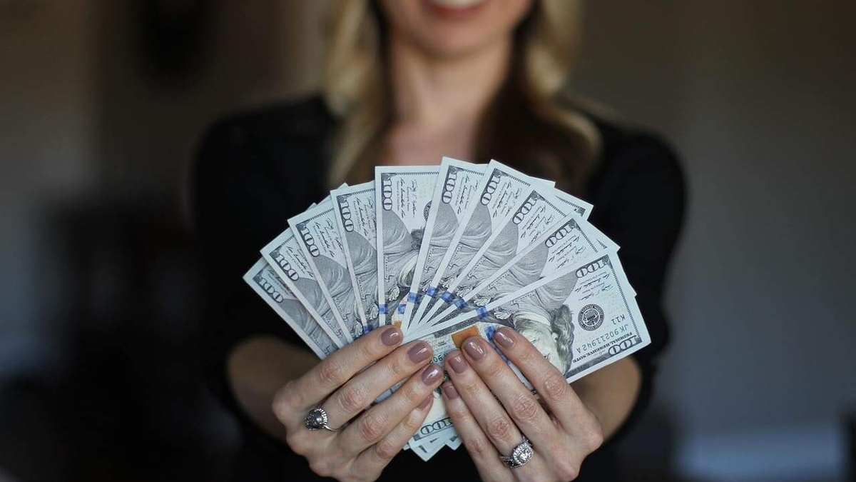 Simple Financial Habits: a woman holding a fanned-out stack of U.S. hundred-dollar bills toward the camera, with her face out of focus in the background.