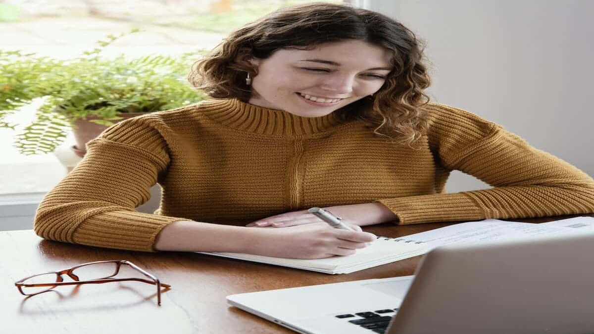 A young woman with curly brown hair, wearing a mustard yellow knitted sweater, is happily writing in a small notebook with a pen. She is seated at a wooden desk with a laptop partially visible and a green potted plant in the background, suggesting she is working or studying from home. She has a warm, focused smile.