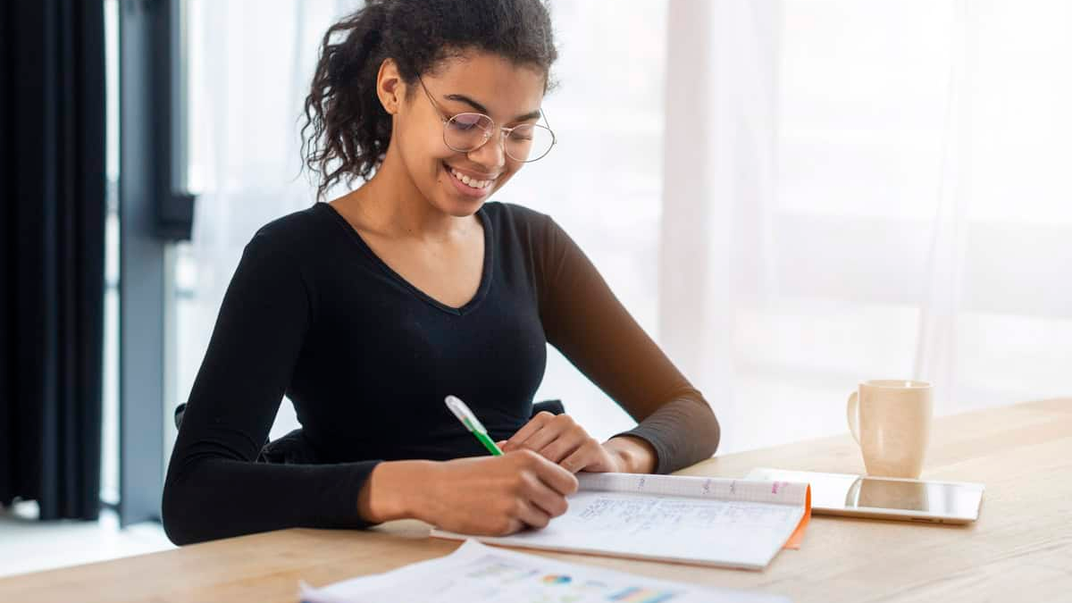 A smiling young woman with dark, curly hair tied back in a ponytail and wearing glasses is happily writing in a notebook with a green pen. She is dressed in a black long-sleeved top and is seated at a brightly lit desk with a window in the background. A mug and a tablet are also visible on the desk.