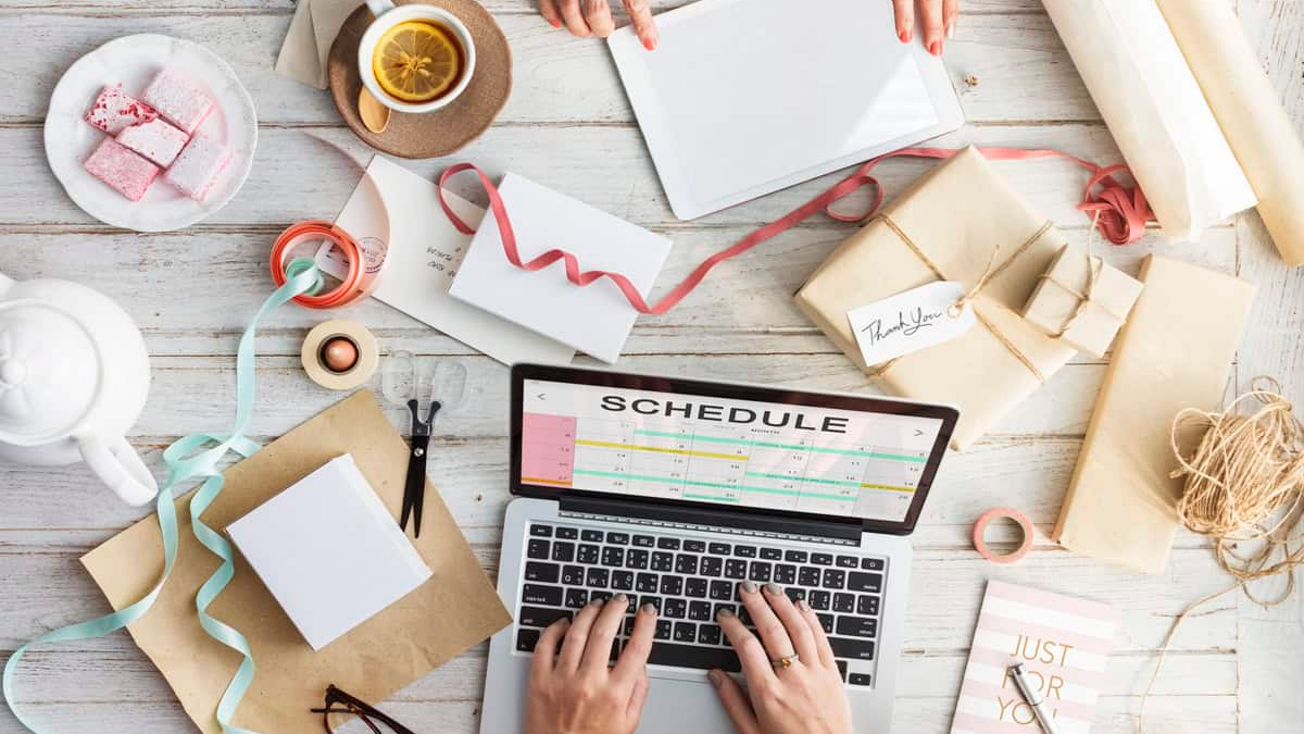 An overhead view of a busy, light-colored wooden desk where two people are collaborating on a project, possibly related to events or a small business. A silver laptop is central, displaying the word "SCHEDULE" on the screen. The desk is cluttered with gifts wrapped in brown paper, ribbons, notebooks, a teapot, a cup of tea, and pink pastries, suggesting planning, crafting, or creative work.