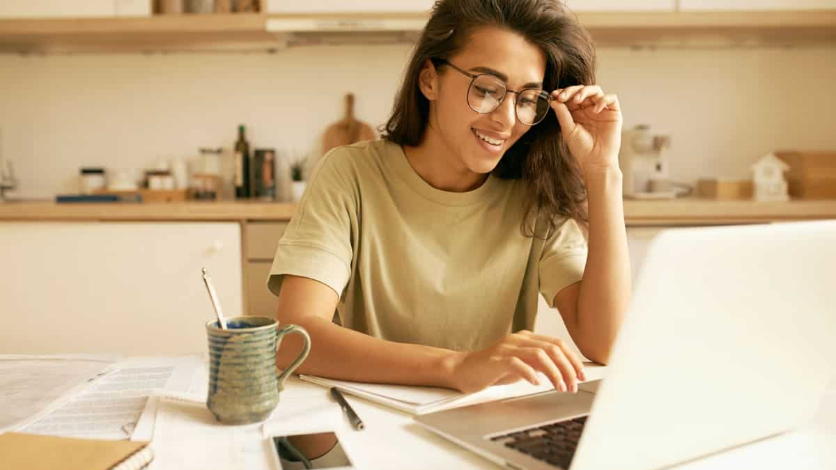 kredit GEHALT Aufteilung The image shows a young woman with dark hair, wearing glasses and a light-colored (possibly olive green) t-shirt, working at a kitchen counter or table.