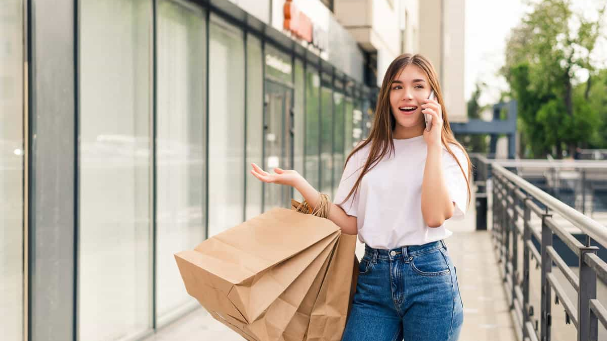 Einkaufen A young woman with long brown hair, wearing a white t-shirt and blue jeans, is walking outdoors past a modern storefront or apartment building. She is talking on a mobile phone, holding several large brown paper shopping bags in one arm, and gesturing with her free hand. She appears excited or engaged in the conversation.