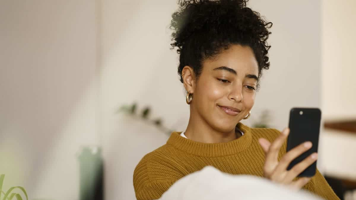 The image features a young woman with curly dark hair pulled up, wearing a mustard yellow or ochre-colored sweater, relaxing while looking at her smartphone.