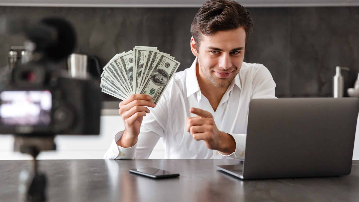 Einkommenspotenzial nachhaltig steigern: strukturierte Strategien für mehr Einkommen ohne Berufswechsel. Earning potential illustrated by a man holding U.S. dollar bills while working on a laptop and recording content with a camera, suggesting online income growth and monetization strategies
