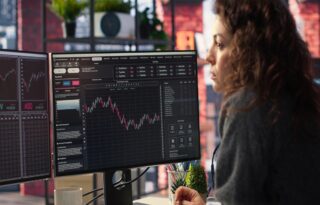 A focused woman with curly brown hair is seated in front of two computer monitors displaying complex financial charts and trading data. Her face is illuminated by the screen, which shows a red and green candlestick chart in the center. She appears to be actively analyzing the stock market or investment performance in an office or home setting.