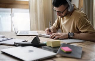 A focused young man wearing glasses and a tan shirt sits at a wooden desk cluttered with notebooks and a laptop. He is wearing over-ear headphones and diligently writing notes in a small spiral notebook with a pen, possibly studying or working from home. Natural light streams in from a window on the left.
