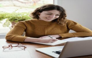 A young woman with curly brown hair, wearing a mustard yellow knitted sweater, is happily writing in a small notebook with a pen. She is seated at a wooden desk with a laptop partially visible and a green potted plant in the background, suggesting she is working or studying from home. She has a warm, focused smile.