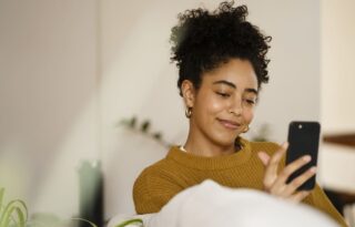 The image features a young woman with curly dark hair pulled up, wearing a mustard yellow or ochre-colored sweater, relaxing while looking at her smartphone.