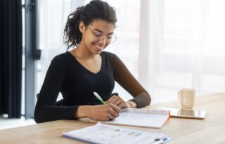 A smiling young woman with dark, curly hair tied back in a ponytail and wearing glasses is happily writing in a notebook with a green pen. She is dressed in a black long-sleeved top and is seated at a brightly lit desk with a window in the background. A mug and a tablet are also visible on the desk.