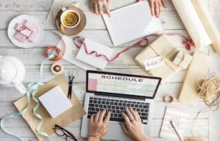 An overhead view of a busy, light-colored wooden desk where two people are collaborating on a project, possibly related to events or a small business. A silver laptop is central, displaying the word 