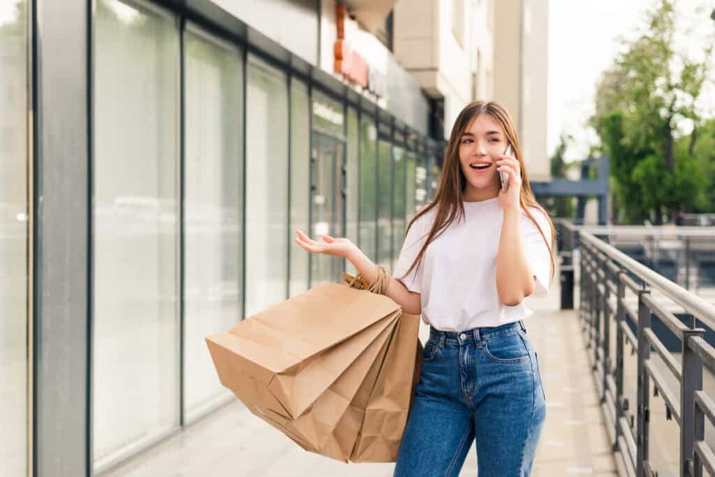 Einkaufen A young woman with long brown hair, wearing a white t-shirt and blue jeans, is walking outdoors past a modern storefront or apartment building. She is talking on a mobile phone, holding several large brown paper shopping bags in one arm, and gesturing with her free hand. She appears excited or engaged in the conversation.