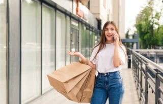A young woman with long brown hair, wearing a white t-shirt and blue jeans, is walking outdoors past a modern storefront or apartment building. She is talking on a mobile phone, holding several large brown paper shopping bags in one arm, and gesturing with her free hand. She appears excited or engaged in the conversation.
