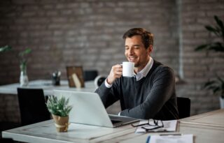 A focused man with glasses and a backpack sits at a wooden desk, diligently writing on a financial planning sheet with colorful sections, surrounded by a laptop