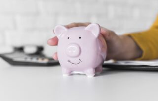 A person wearing a yellow sleeve gently holds a pink ceramic piggy bank with their hand, resting it on a clean white desk or surface. In the background, a calculator and some documents are visible, suggesting the context of budgeting or saving money.