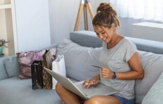 The image shows a young woman comfortably seated on a sofa, happily engaged in online shopping using a laptop and a credit card.