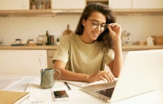 The image shows a young woman with dark hair, wearing glasses and a light-colored (possibly olive green) t-shirt, working at a kitchen counter or table.
