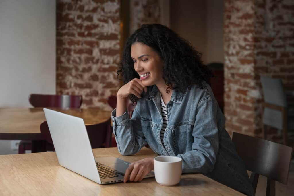 Finanzpuffer schufa The image features a young woman smiling warmly while engaging with a laptop in a cozy, brick-walled setting, likely a cafe or a co-working space.