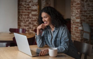 The image features a young woman smiling warmly while engaging with a laptop in a cozy, brick-walled setting, likely a cafe or a co-working space.