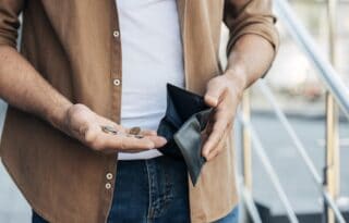 The image is a close-up of a person, likely a man, holding an empty black bifold wallet in one hand and a few small coins in the other.