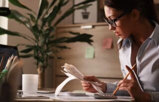 The image shows a woman with dark hair and glasses working intently at a desk, seemingly managing finances or reviewing documents.