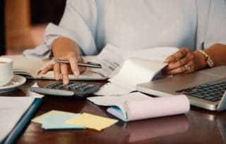 A close-up view of a person's hands diligently working on financial tasks at a desk. One hand holds a pen over a paper receipt or document, while the other hand rests near a calculator. A laptop, a notebook, and a coffee cup are also visible on the desk, suggesting the person is reviewing bills, calculating budgets, or handling finances.