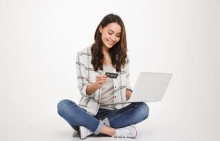 The image is a studio shot of a young woman sitting cross-legged on the floor, using a laptop and a credit card, likely for an online transaction.