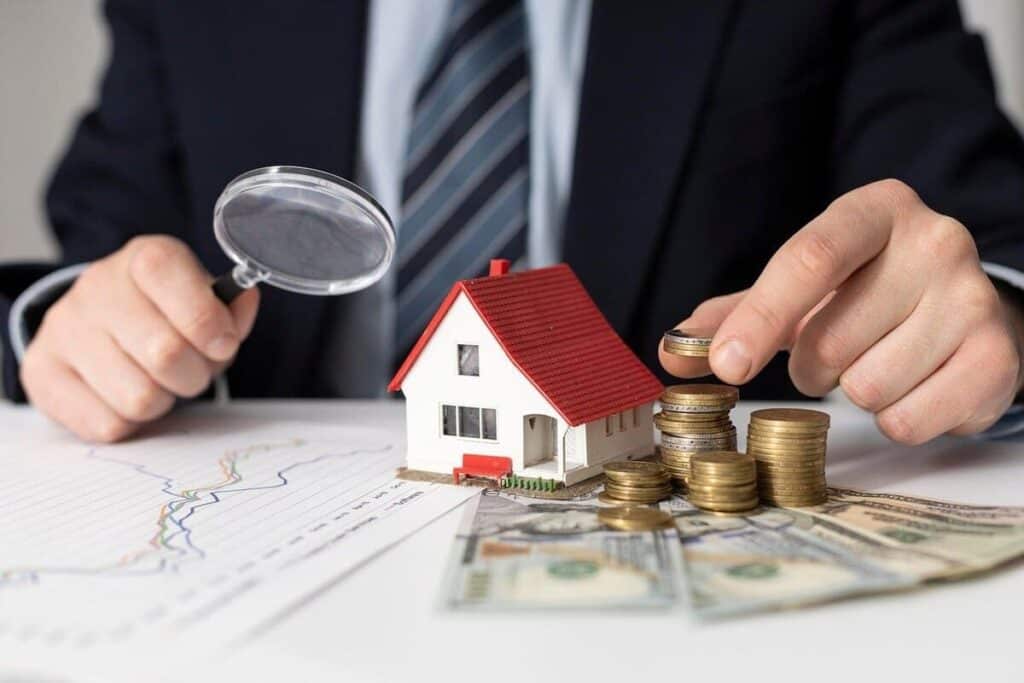 A person in a suit examining a small model house with a magnifying glass, surrounded by stacks of coins, dollar bills, and financial charts on the table.