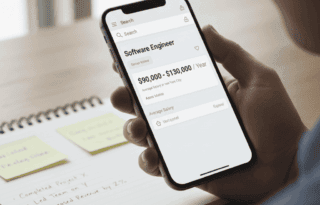 A hand holding a smartphone displaying a search result from a salary aggregation website, showing a clear average salary range for a specific job title. In the background, blurred notes on a desk with bullet points of achievements are visible.