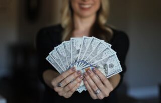 Simple Financial Habits: a woman holding a fanned-out stack of U.S. hundred-dollar bills toward the camera, with her face out of focus in the background.