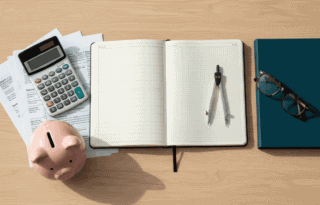 A middle-aged man sitting at a dimly lit kitchen table at night, looking stressed while working on a laptop and writing in a notebook filled with financial calculations.