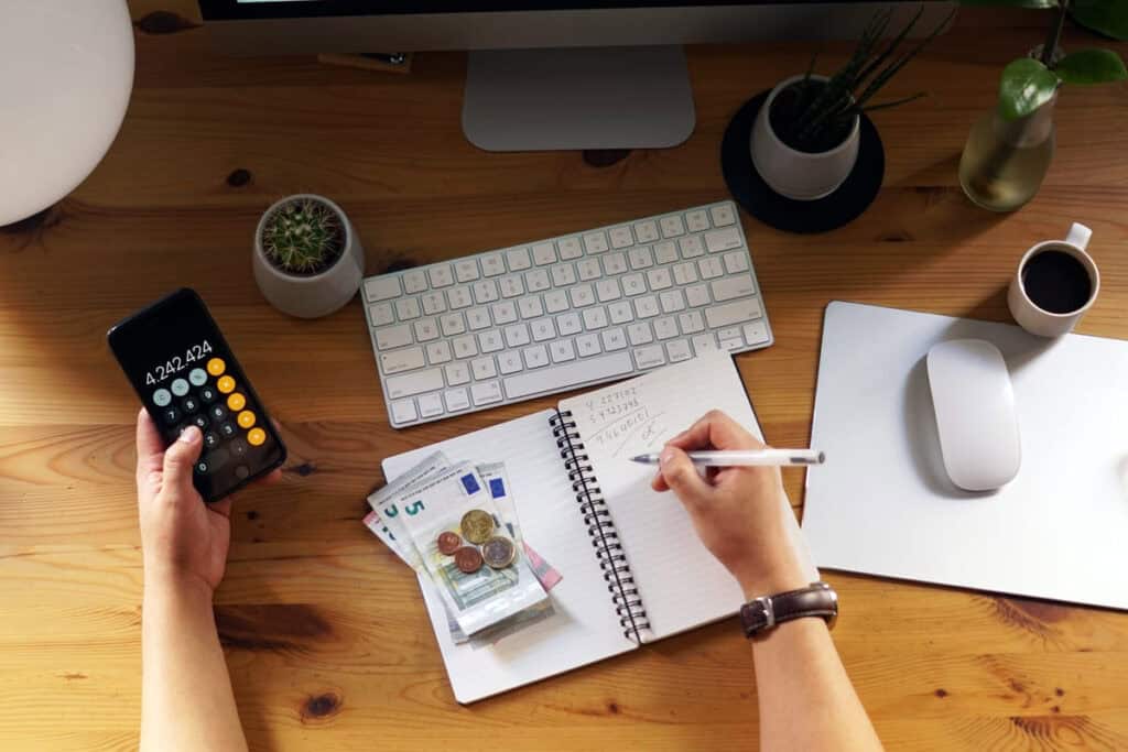 personal finance planning workspace with a person writing in a notebook beside a calculator, keyboard, coffee cup, and desk accessories