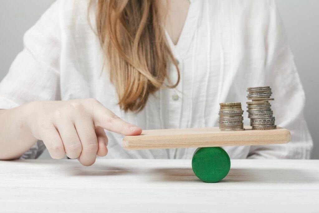 Spend less by keeping financial balance: a person adjusting a wooden scale with stacked coins on one side.