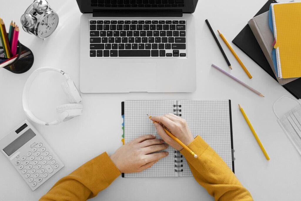 Wealth planning workspace with a laptop, notebook, calculator, pencils, and hands writing financial notes on a clean desk