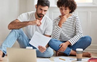 ETFs vs Stocks couple sitting on the floor at home reviewing financial documents and charts, with a laptop and coffee cups nearby, discussing investment options and planning their finances together.