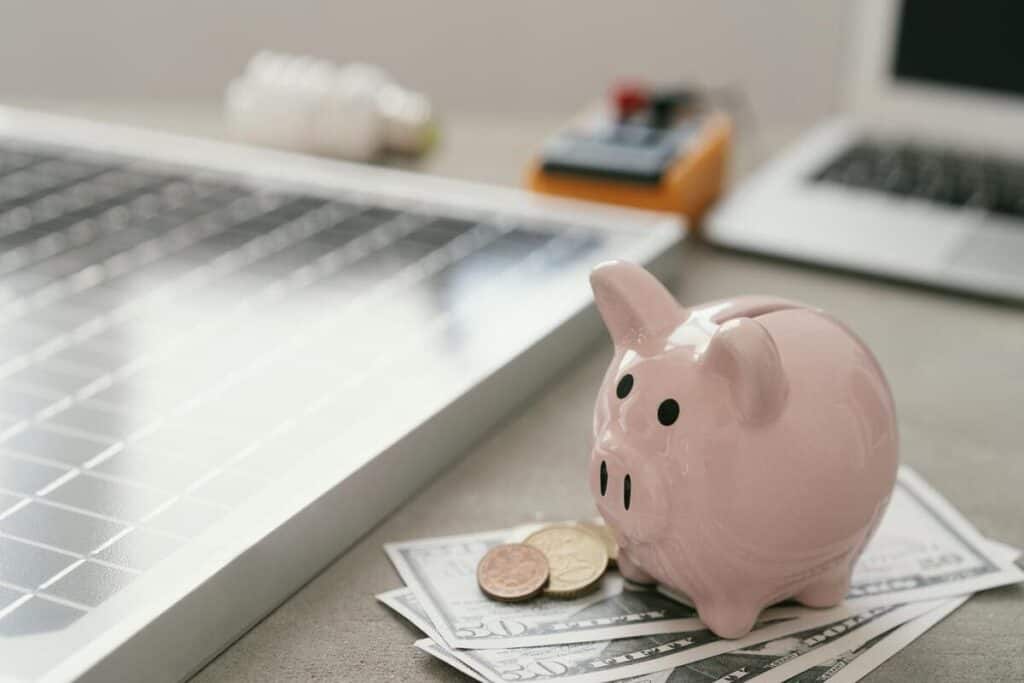 checking vs savings account concept shown by a pink piggy bank sitting on dollar bills next to a laptop and coins on a desk
